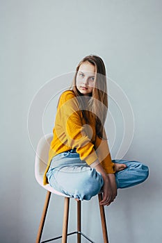 Teenage girl looks at camera.sitting on white modern white chair with two leg raise up. Beautiful girl in yellow sweater and blue