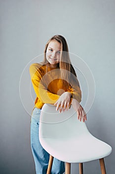 Teenage girl looks at camera standing lean on white modern chair. Beautiful girl in yellow sweater and blue jeans in home interior
