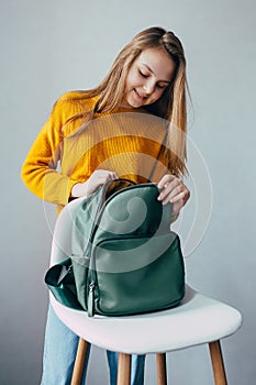 Teenage girl looks inside leather rucksacks that lies on a white modern chair. Beautiful girl in yellow sweater and blue jeans sta