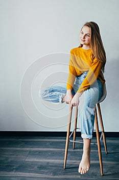 Teenage girl looks at left side.sitting on white modern chair with one  leg raise up. Beautiful girl in yellow sweater and blue je