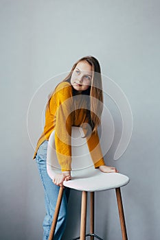 Teenage girl looks at left side standing lean on white modern white chair. Beautiful girl in yellow sweater and blue jeans in home