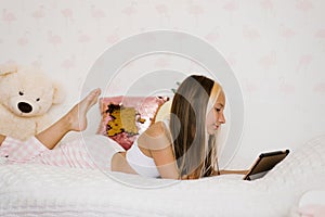 Teenage girl lying on the bed in her pajamas in the morning is watching educational programs on a tablet
