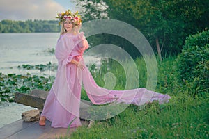 A teenage girl in a pink dress and a wreath of flowers on her head stands by the river. Summer. Nature. Inspiration