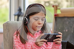 A teenage girl is playing on a smartphone with headphones with a microphone, sitting in pajamas on a blurry neutral background. Co