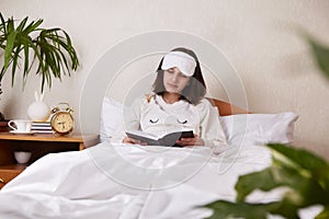 A teenage girl is reading a book in bed under a blanket.