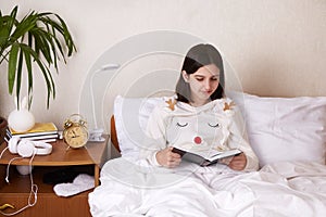 A teenage girl is reading a book in bed under a blanket.