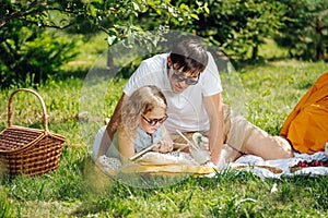 Teenage girl reading book with her father on grass during family picnic