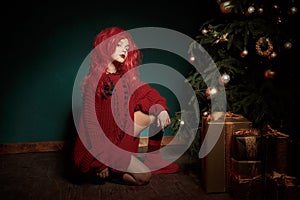 A teenage girl in a red knitted sweater and wig sits on the floor near a Christmas tree and presents. Fashion New Year