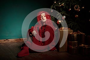 A teenage girl in a red knitted sweater and wig sits on the floor near a Christmas tree and presents. Fashion New Year
