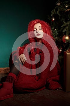 A teenage girl in a red knitted sweater and wig sits on the floor near a Christmas tree and presents. Fashion New Year