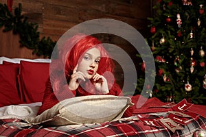 A teenage girl in a red wig lies in bed in a room for Christmas. Fashion, New Year