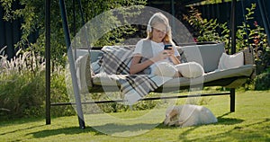 A teenage girl is resting on a garden swing, using a smartphone. Her puppy sits next to her