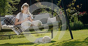 A teenage girl is resting on a garden swing, using a smartphone. Her puppy sits next to her