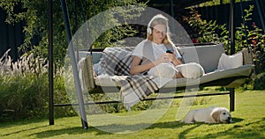 A teenage girl is resting on a garden swing, using a smartphone. Her puppy sits next to her