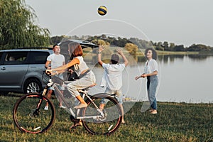 Teenage Girl Riding Bicycle While Mother and Father Playing Volleyball with Her Brother Outdoors by the Lake, Happy Four