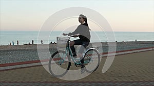 Teenage girl is riding a bike along gravel beach in evening near sea, side view