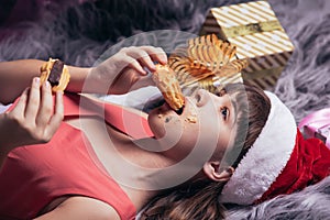 A teenage girl in a Santa hat eats Christmas cakes lying on the carpet