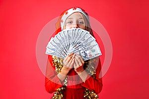 Teenage girl, in Santa`s hat and with tinsel on her neck, with dollars in her hands , isolated on a red background