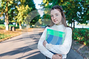 Teenage girl schoolgirl, teenager white sweater summer city, background trees road, warm sweater, hands notebooks books