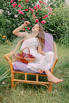 A teenage girl sits on an armchair and eats a cherry