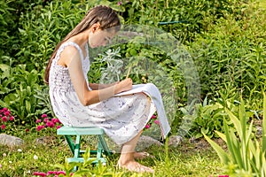 A teenage girl sits on a bench in the garden for her favorite pastime, drawing with a pencil in the album.