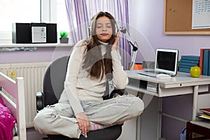 A teenage girl sits cross-legged in a chair and listens to music on headphones.