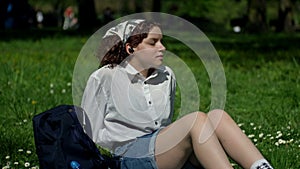 Teenage girl sits on the green grass in the park and listens to music, eyes closed, through earphone.