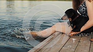 A teenage girl sits pier by the river with a dog of the breed dachshund and splashes bare feet in the water. View from