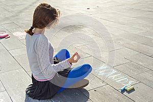 Teenage girl sits in pose meditation, practicing yoga. On the asphalt text yoga kids, written by colored crayons