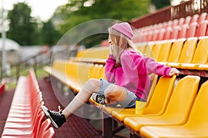 A teenage girl is sitting at the stadium at halftime and talking on the phone with friends. Side view