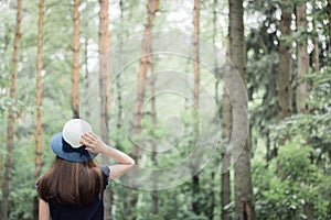 A teenage girl standing with her back holds her hat with her hand in front of tall trees in the forest
