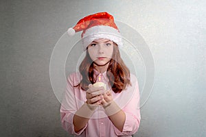 A teenage girl stands at home with a Santa hat and holds a burning white candle in her hands