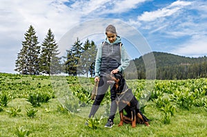 Teenage girl stands with leash in hands next to dog of Rottweiler breed in meadow, against background of fir trees