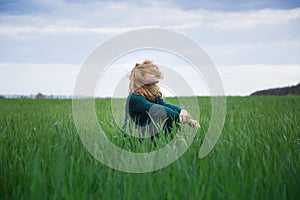 Teenage girl in sweater and long emerald-colored skirt sits on ground among field of young grass