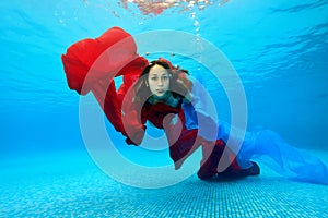 Teenage girl swims underwater in the pool on a blue background and looks at the camera. Portrait. Shooting under water