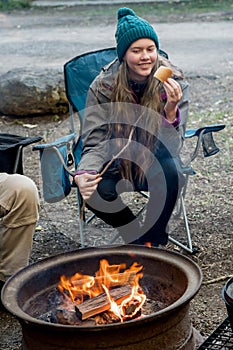 Teenage girl wearing beanie hat eating large marshmallow on a stick roasted over the campfire firepit. Camping family fun