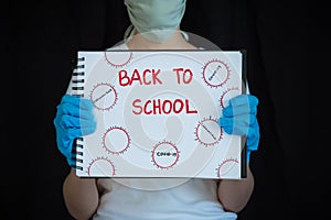 Teenage girl wearing face mask and surgical gloves, holding up notebook that says Back to School