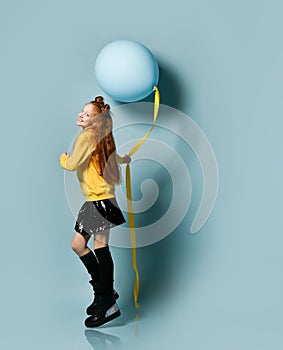 Teenage girl in yellow sweatshirt, black skirt, knee-highs, boots. She holding balloon, posing sideways on blue background.