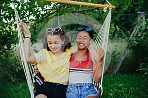 Teenage girls friends outdoors in garden, swinging on swing, having fun, talking during warm summer evening.