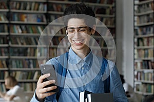 Teenage guy in glasses using smartphone standing in university library