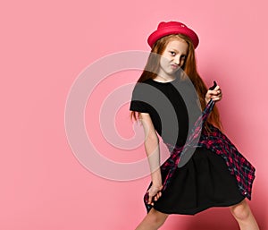 Teenage kid in black dress and red hat. She tightens checkered shirt on her waist while posing against pink background. Close up