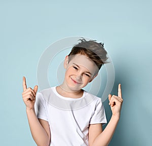 Teenage kid in white t-shirt. He smiling, raised his forefingers up, posing against blue studio background