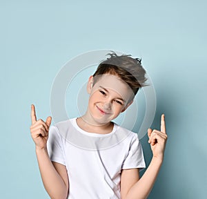 Teenage kid in white t-shirt. He smiling, raised his forefingers up, posing against blue studio background photo