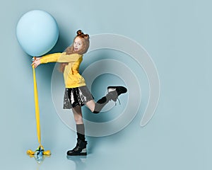 Teenage kid in yellow sweatshirt, black skirt, knee-highs, boots. Smiling, holding balloon, posing sideways on blue background.