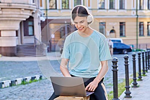 Teenage male student in headphones using laptop outdoor