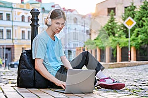 Teenage male student in headphones using laptop outdoor