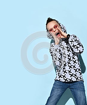 Teenage male in sunglasses and hood, blue jeans and stylish hoodie, black bracelet. Calling someone, posing on blue background