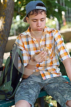 a teenage school boy sits on a bench in a city park and eats sandwiches, a bright summer day
