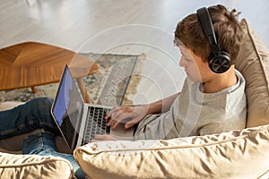 A teenage , sits on a sofa in a room, wearing black headphones on his head, plays a computer game on a laptop.