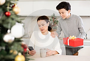 Teenage son pleases mom with box of Christmas gifts next to Christmas tree
